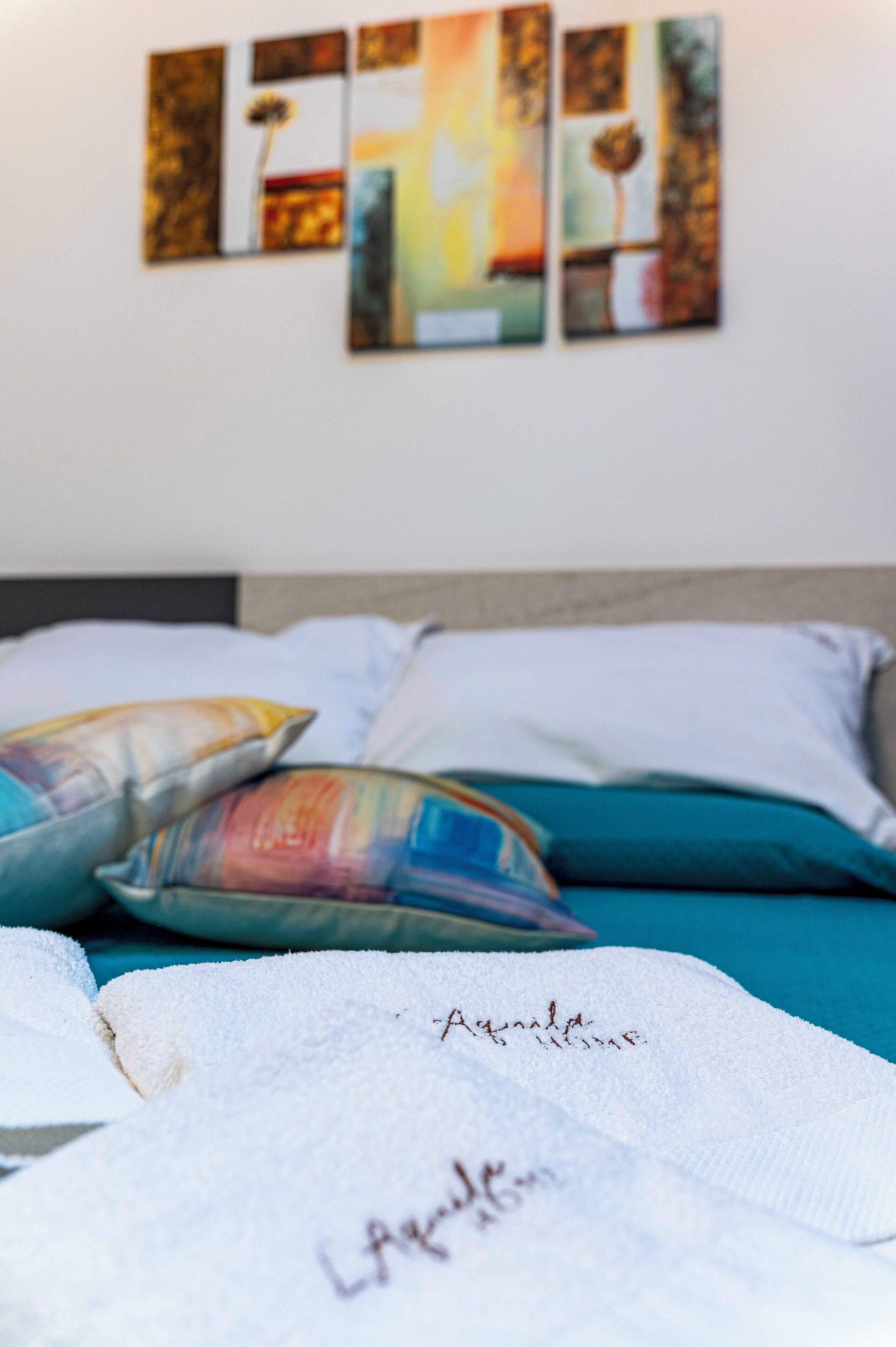 Un letto con coperta e cuscini bianchi, una testata e un muro decorato con arte astratta.