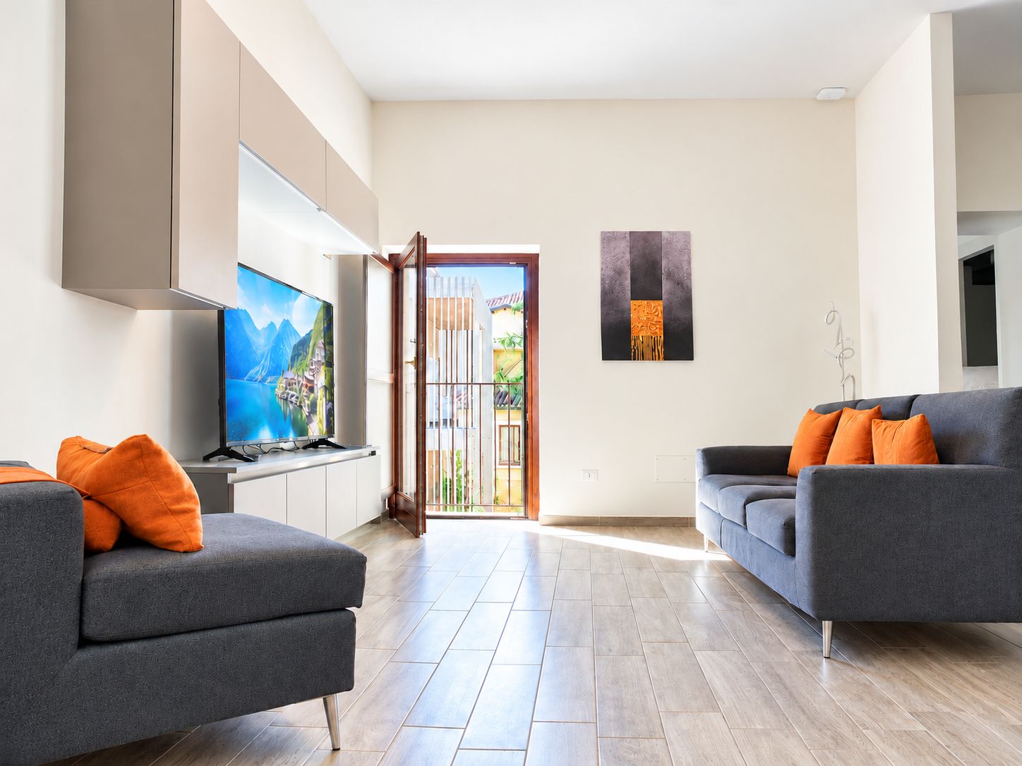 Una sala moderna con un televisor de pantalla plana, dos sofás grises con cojines naranjas, piso de madera y un ventanal con vista a un edificio y follaje.