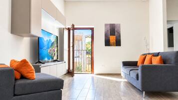 Una sala moderna con un televisor de pantalla plana, dos sofás grises con cojines naranjas, piso de madera y un ventanal con vista a un edificio y follaje.
