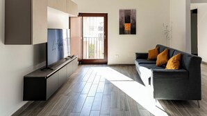 Een moderne woonkamer met een flatscreen-televisie, een grijze bank met oranje kussens en een groot raam.
