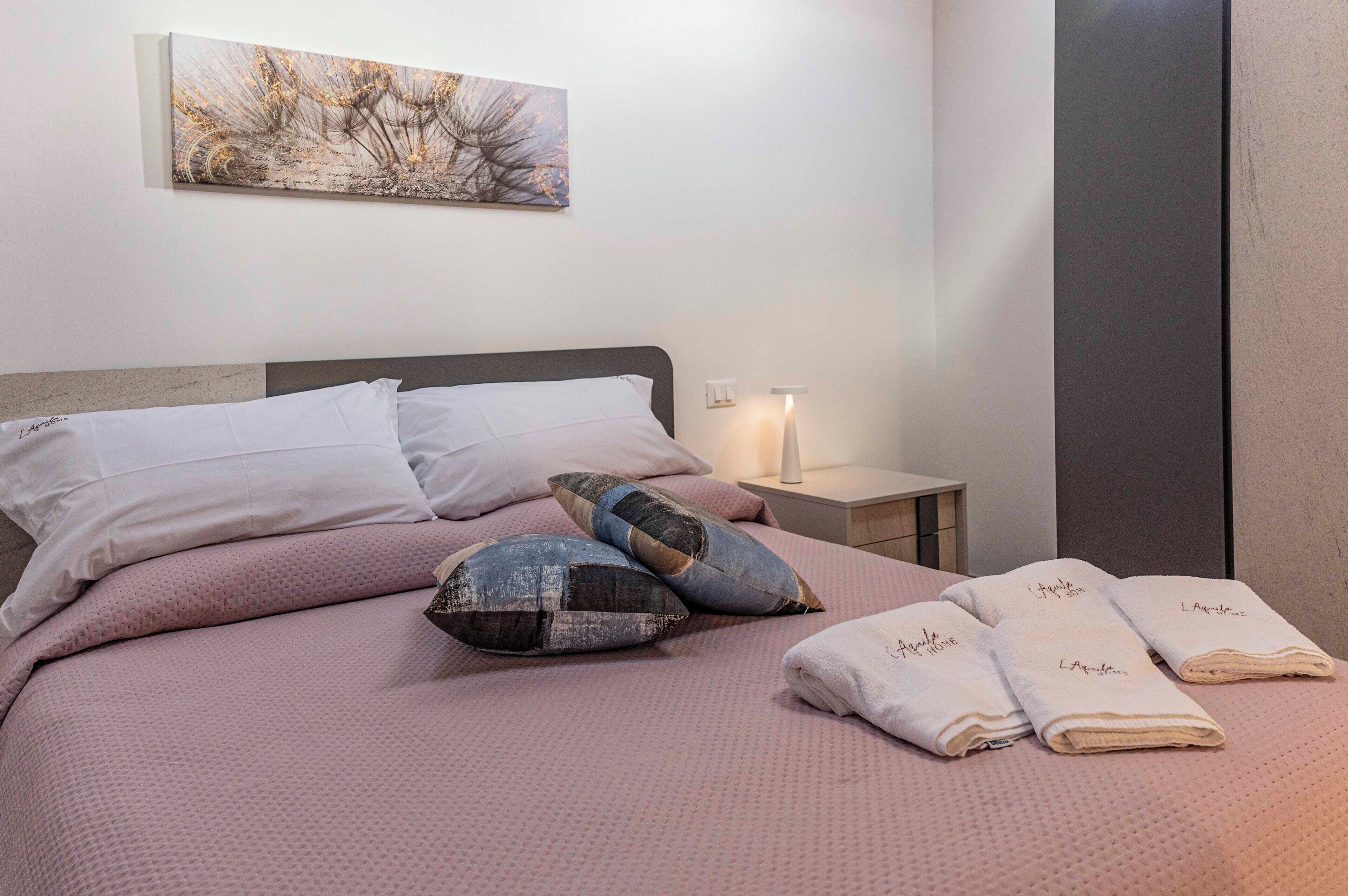 Un lit bien fait, avec une couvre-lit rose, deux oreillers et trois serviettes pliées, chacune portant une inscription.
