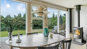 Un espace de repas moderne avec une table ronde, des chaises, un luminaire suspendu, une vue dégagée grâce à de grandes fenêtres et un poêle à bois.