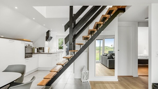 A modern kitchen with white cabinets, a dining area with a table and chairs, and a staircase with wooden steps and black railings. - "Hejdie" - 150m from the sea by Interhome (Hals)