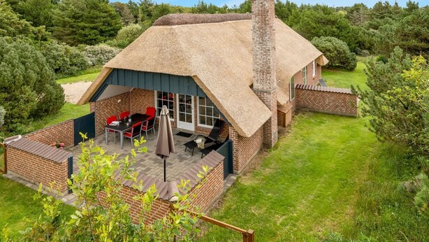 Una cabaña con techo de paja, una chimenea de ladrillo, un patio con mobiliario exterior y un jardín exuberante.