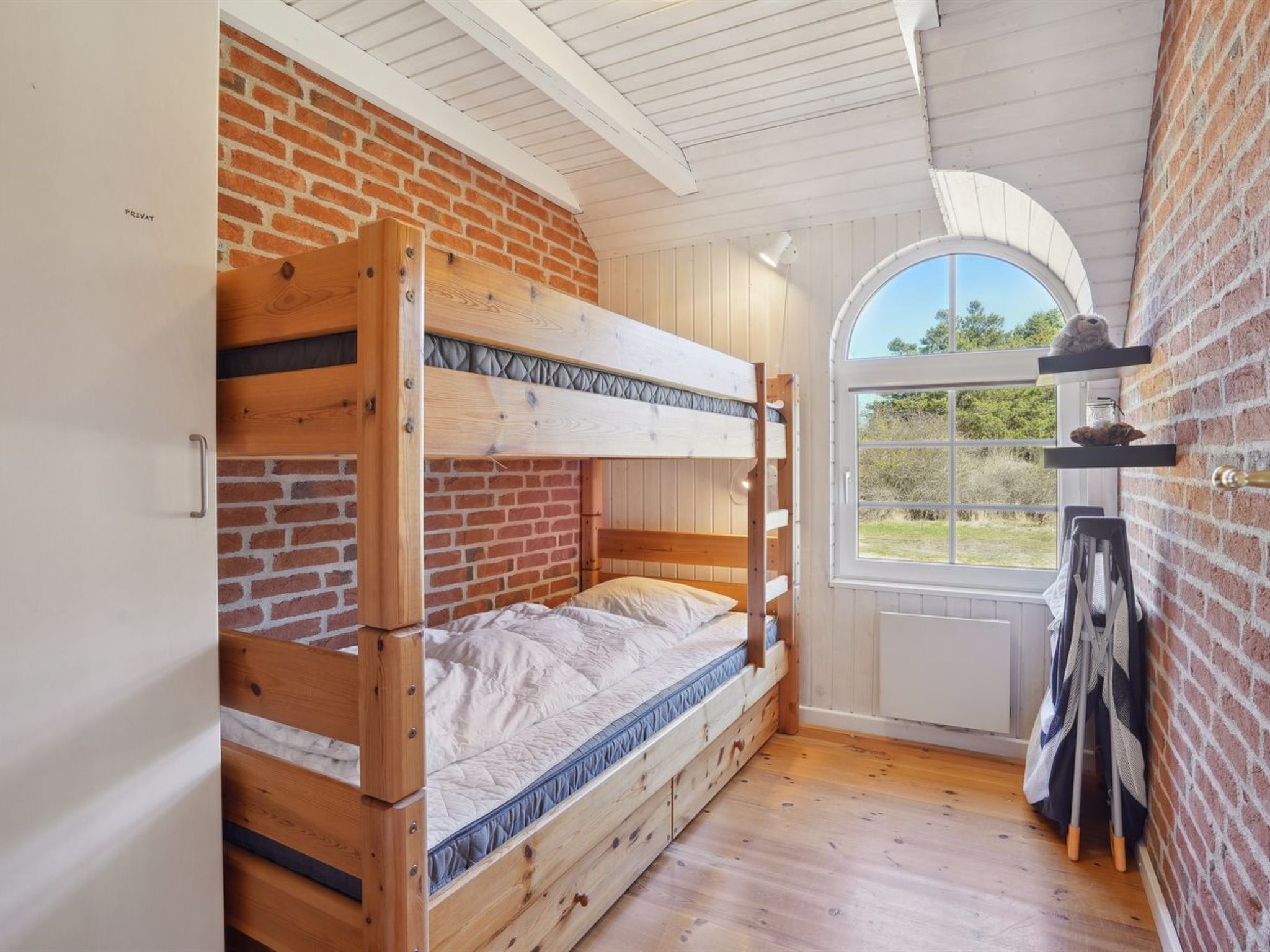 Quarto com beliche, parede de tijolos e janela com vista para um jardim.