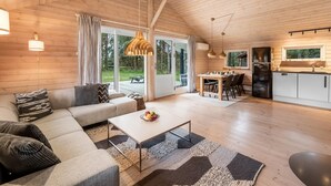 A spacious living area with a sofa, coffee table, and dining table. There is a kitchen area with a refrigerator and a window overlooking a deck.