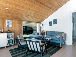 Una sala de estar moderna con techo de madera, un sofá azul, una silla negra, una mesita con una lámpara y vistas al exterior.