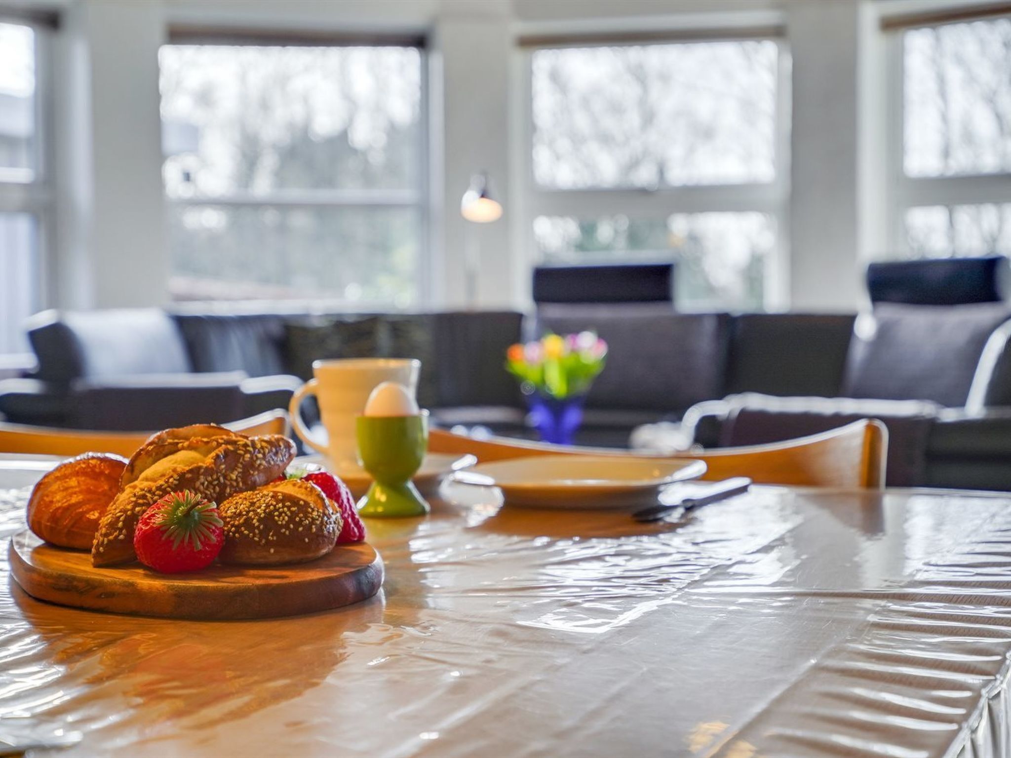 Ein Frühstückstisch mit Brot, Erdbeeren und einer Tasse in einem modernen Zimmer mit großen Fenstern und einer Couch.