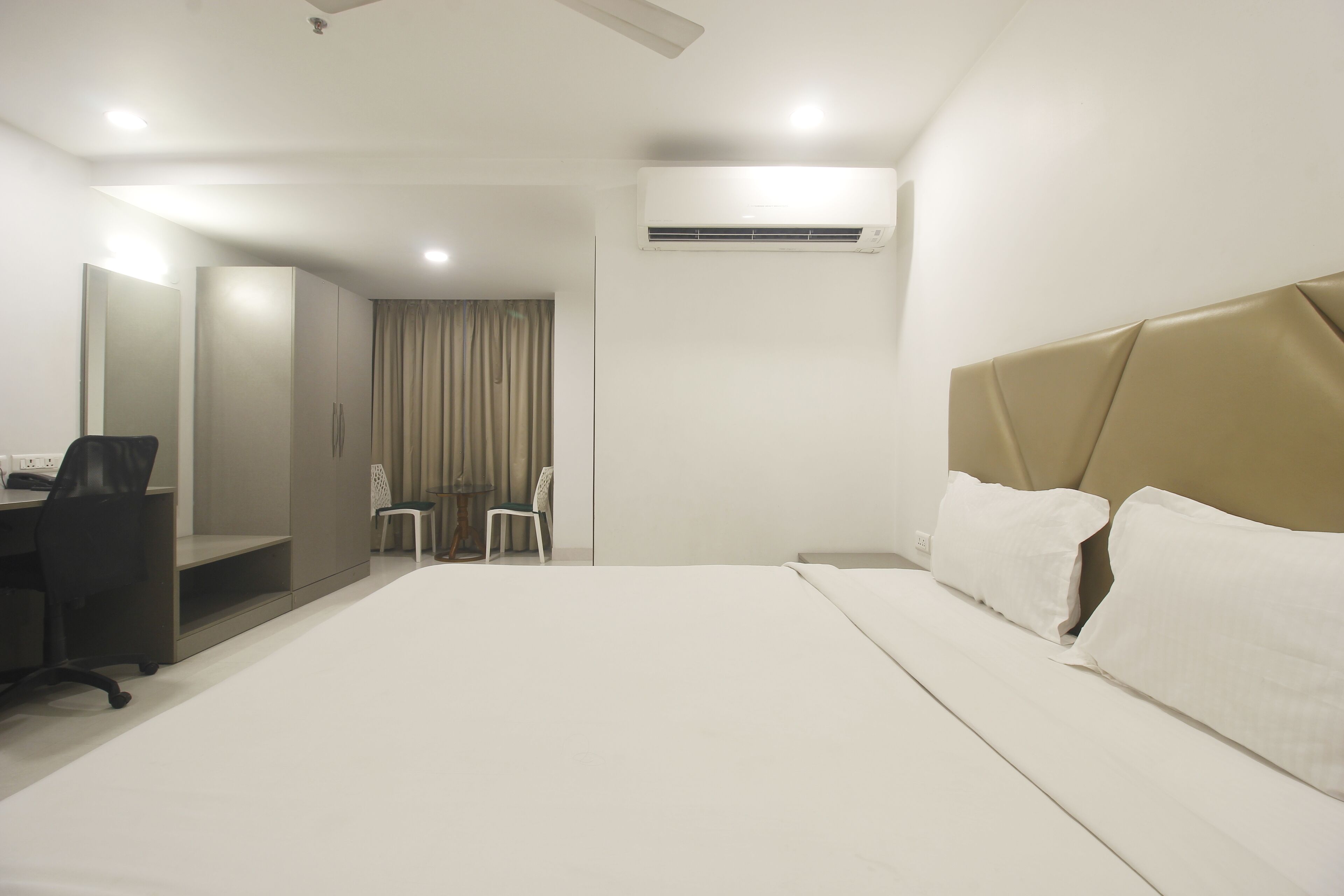 A hotel room with a large bed, a desk, two chairs, and an air conditioning unit.