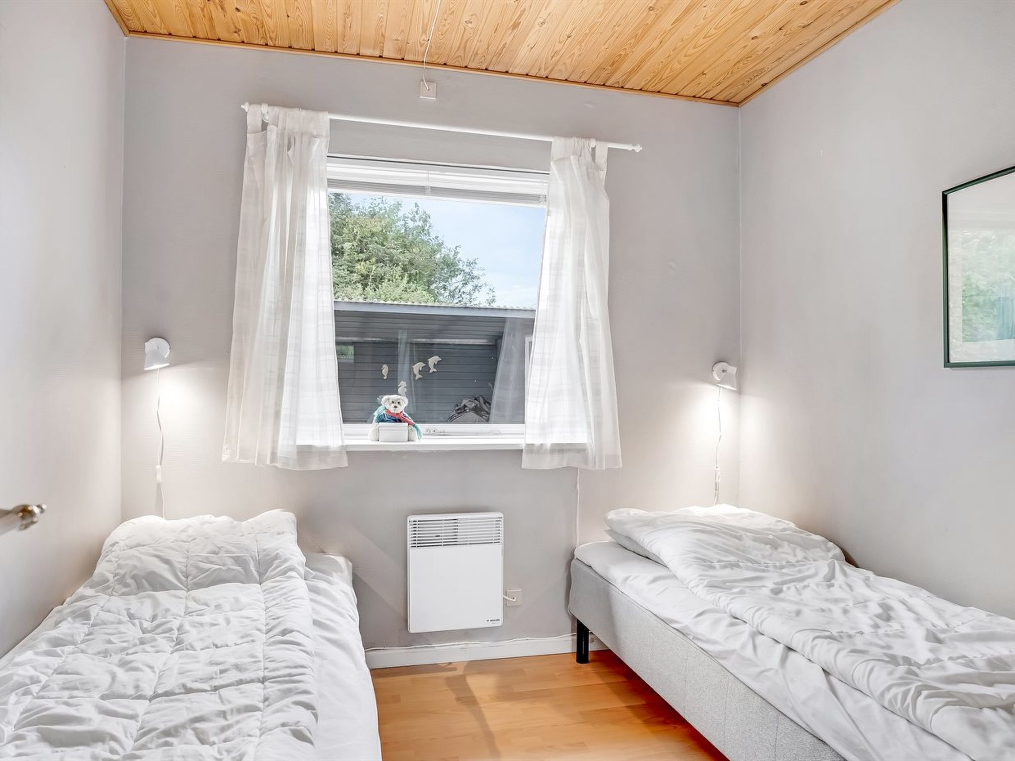 A bedroom with two single beds, a window with white curtains, a wall-mounted heater, and a framed picture on the wall.