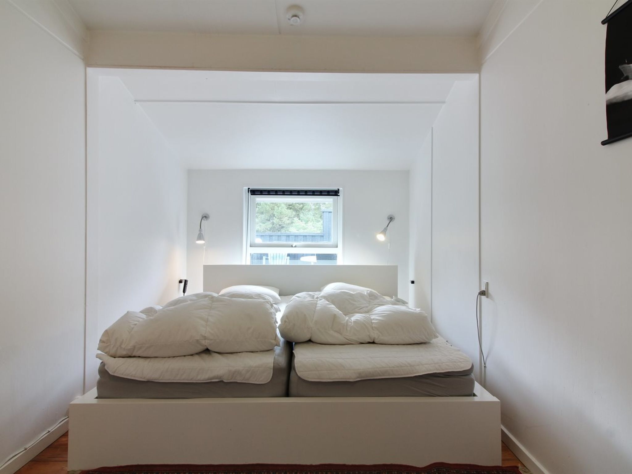 A narrow, white-walled room with a bed centered under a window.