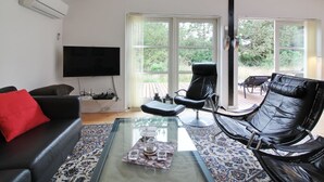 A modern living room with a black leather sofa, a red pillow, a glass coffee table, and a black leather chair. There is a flat-screen TV mounted on the wall and a large window with a view of greenery outside. - "Nienke" - 700m from the sea by Interhome (Løkken)