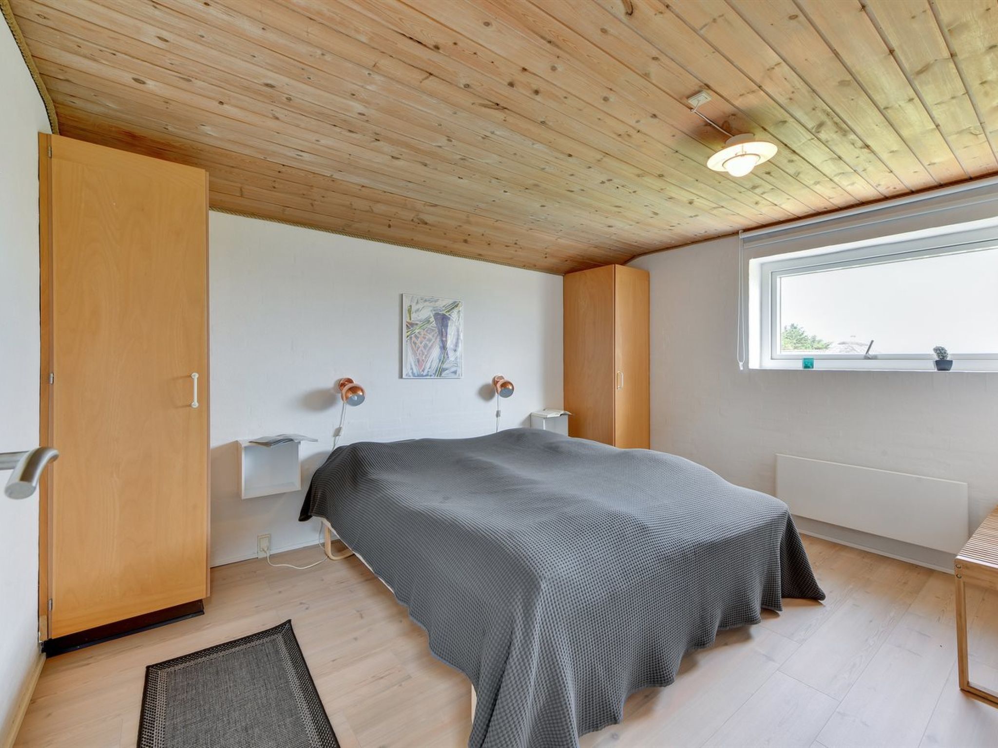 A bedroom with a wooden ceiling, a bed with a grey bedspread, a wooden wardrobe, a bedside table with lamps, a window with a view, and a small potted plant on the windowsill.