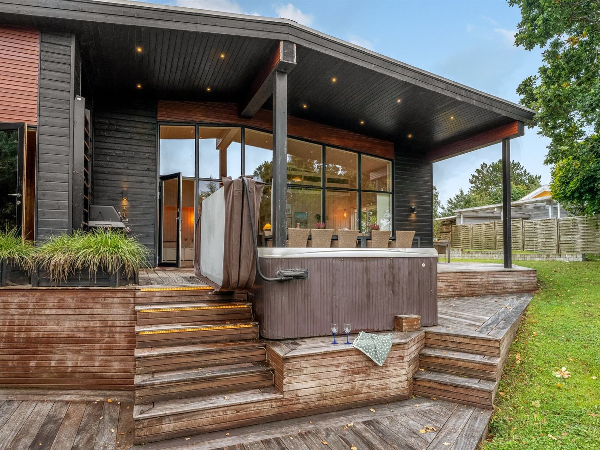 A modern outdoor hot tub area with wooden steps leading up to it, surrounded by a deck and a covered seating area.