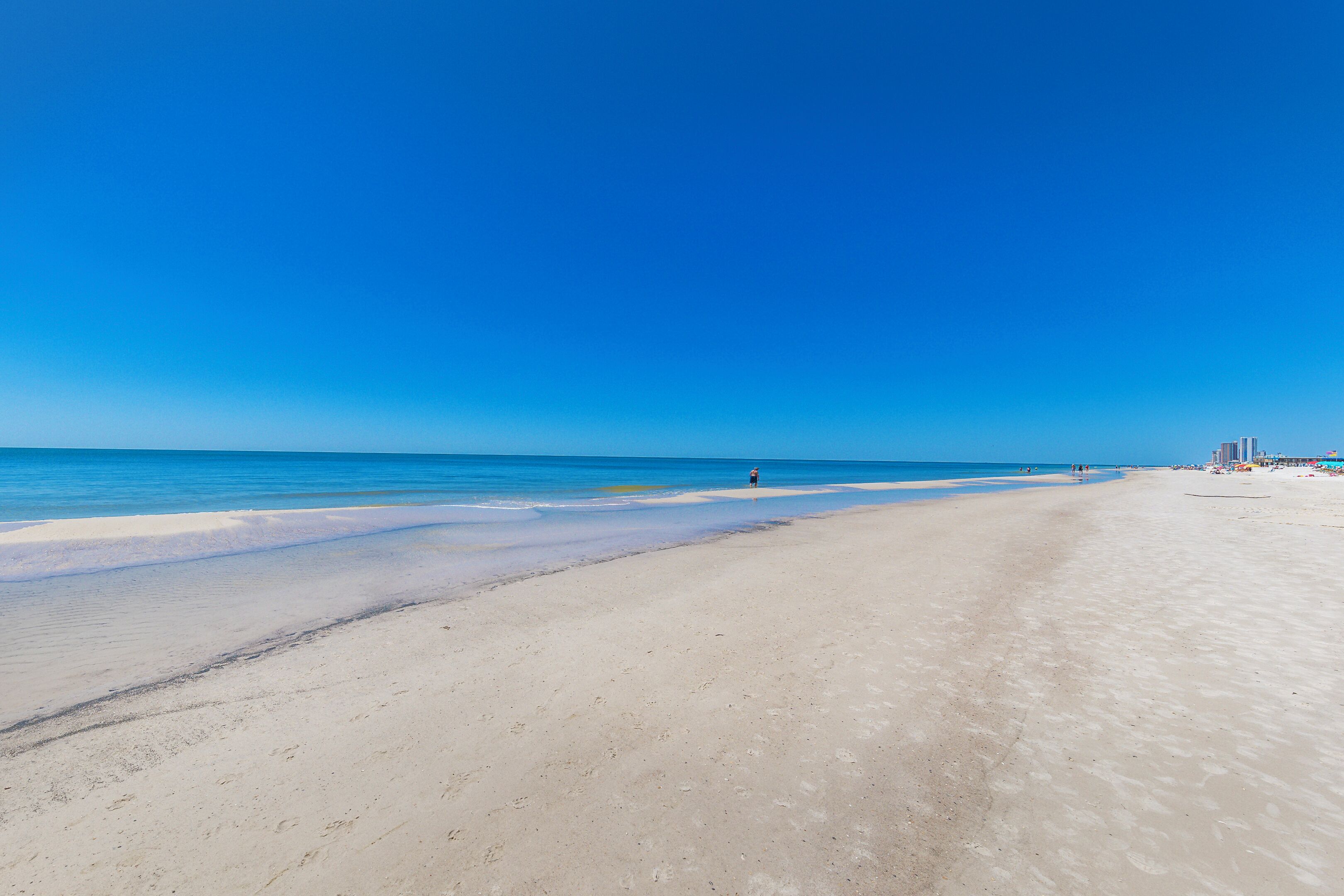 Ein weiter Sandstrand mit klarem, blauem Wasser und ein paar Personen in der Ferne.