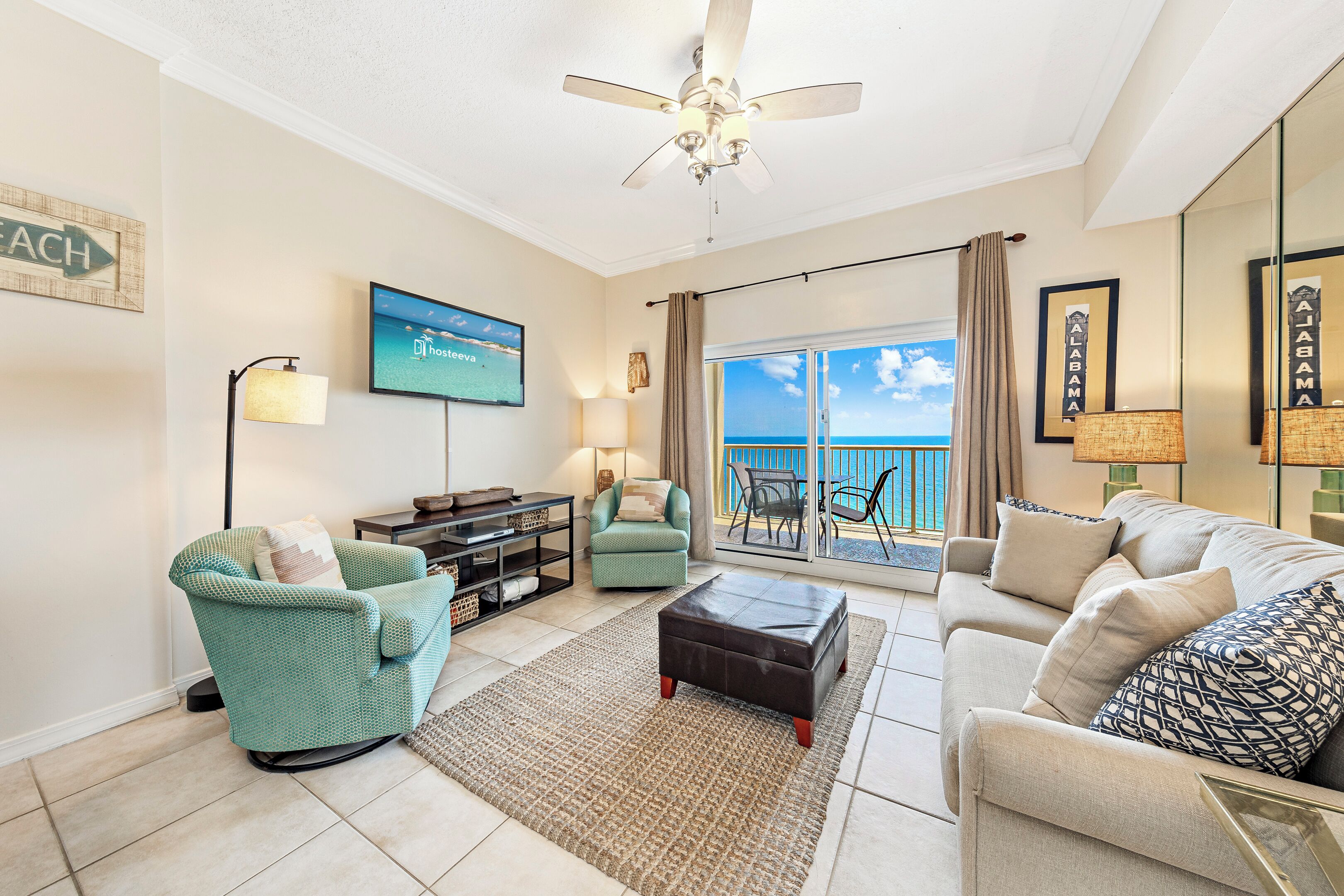 A spacious living room with a sofa, armchair, and ottoman, a television, and a large window with a view of the ocean.