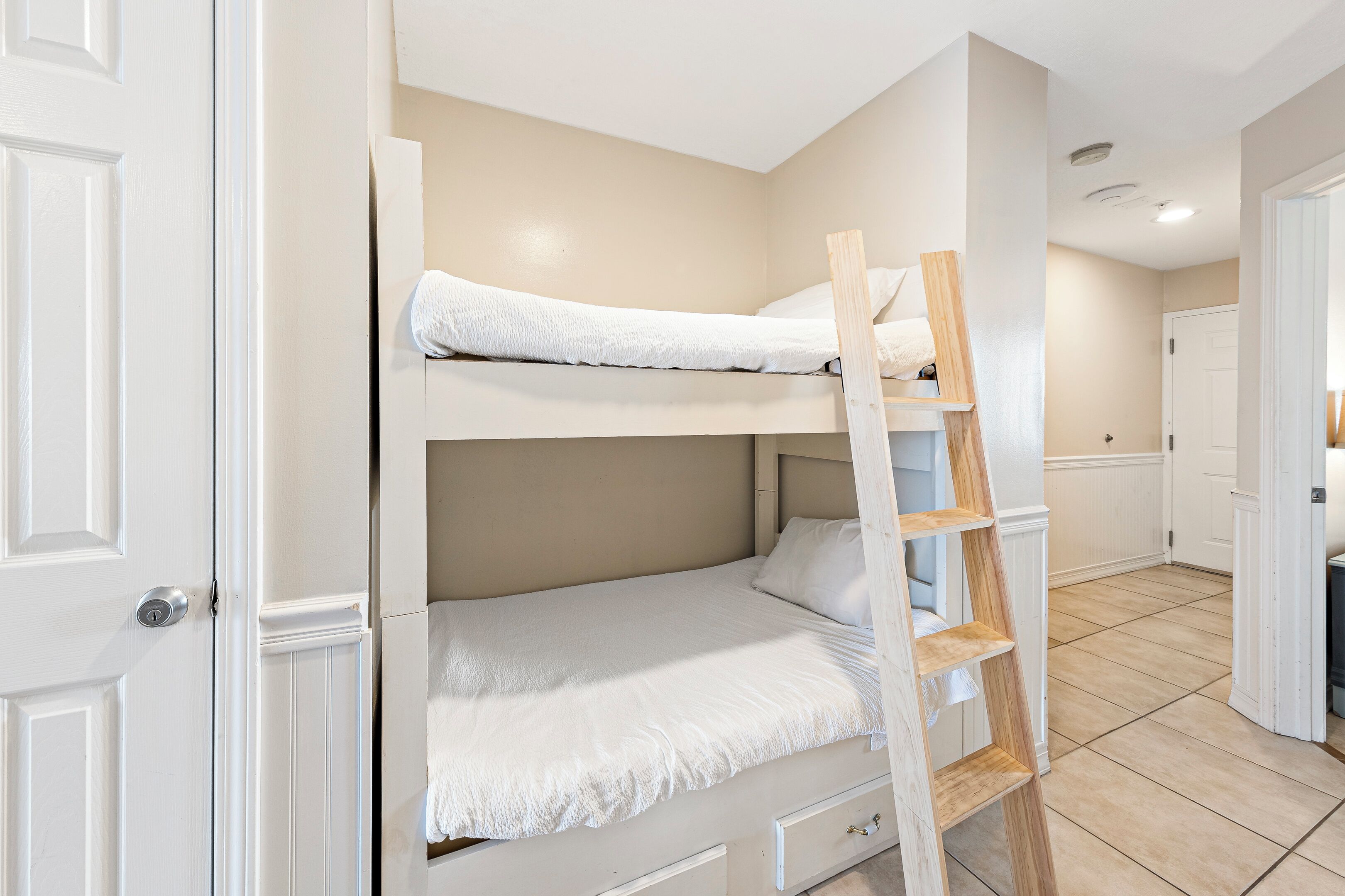 Un lit superposé avec une échelle en bois, une porte blanche et un sol carrelé.