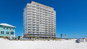 Et hotel med flere etager og altaner, beliggende ved en strand med en klar blå himmel.