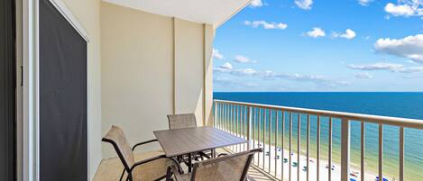 Un balcon avec une table et des chaises, donnant sur une plage parsemée de parasols et sur l’océan.