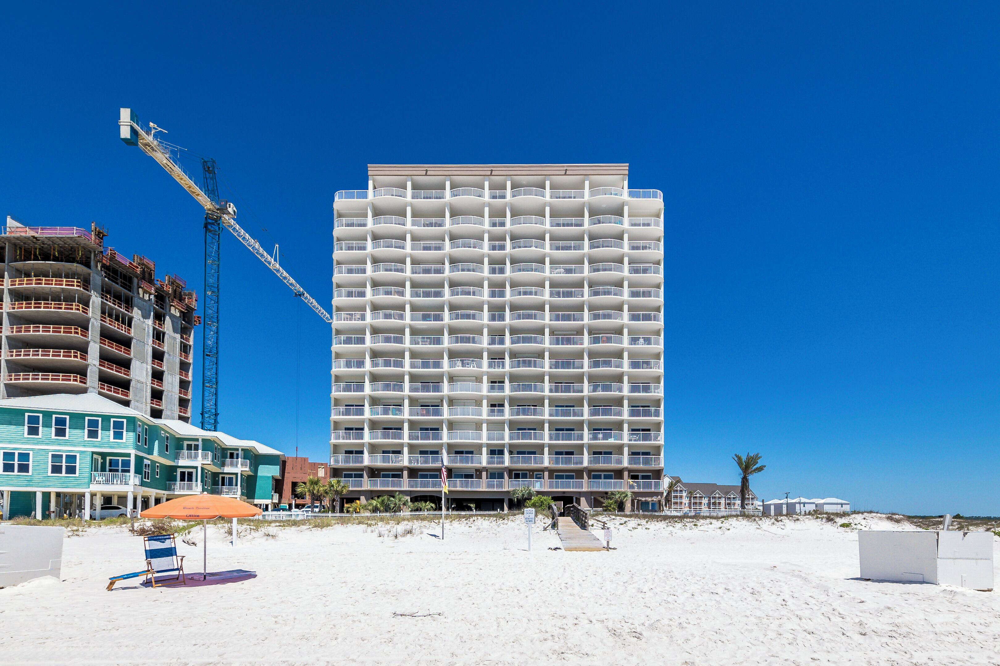 A multi-story hotel with a balcony on each floor, located on a beach with a clear blue sky.