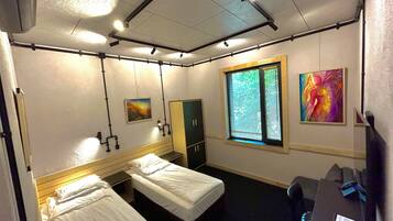 Habitación de hotel con dos camas individuales, un escritorio con silla, armario, ventana con persianas y dos lámparas de pared.