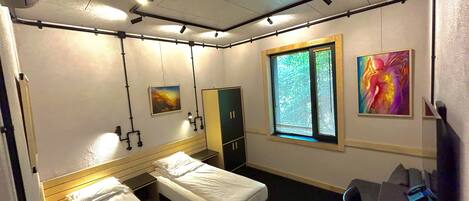 A hotel room with two single beds, a desk with a chair, a wardrobe, a window with blinds, and two wall-mounted light fixtures.