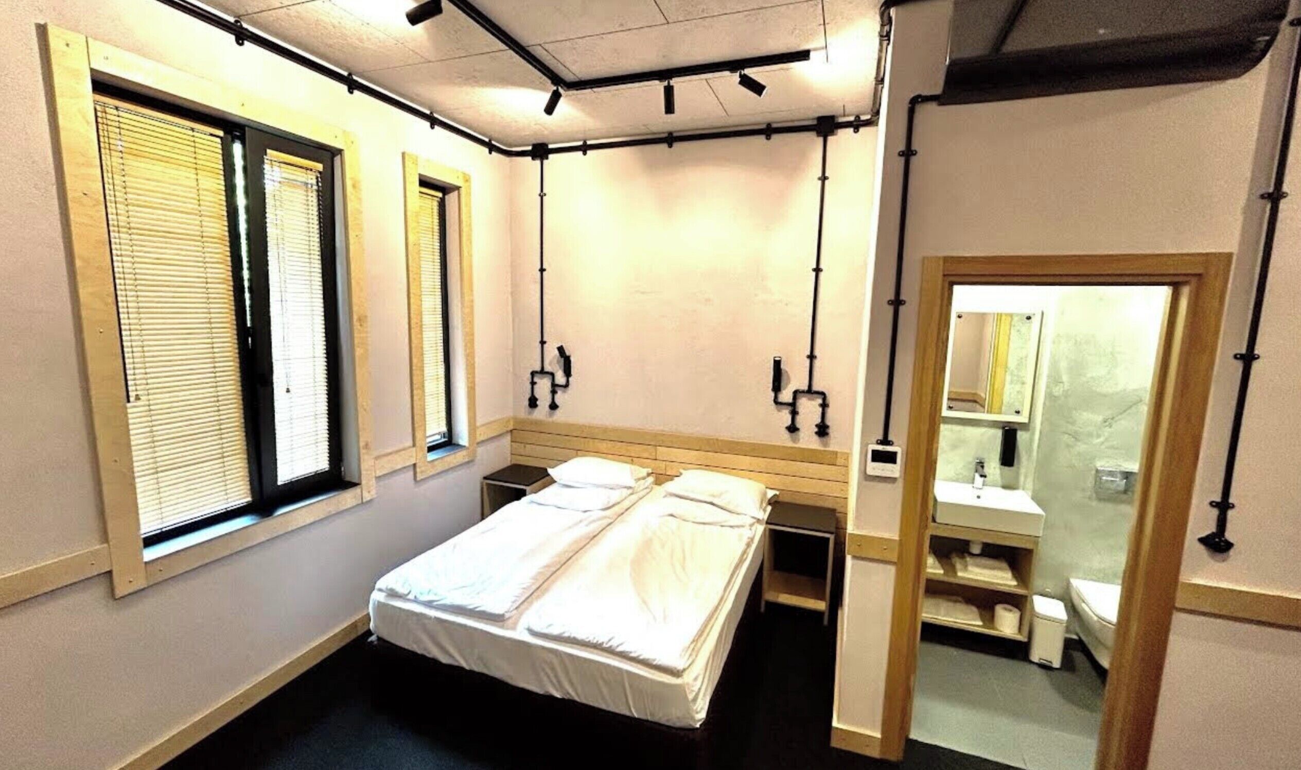 Una habitación pequeña con una cama individual, cabecera de madera, mesita de noche, ventana con persianas, espejo y un inodoro.