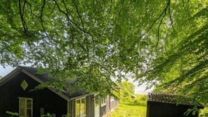 Uma cabana rústica com telhado inclinado, um pequeno alpendre e um galpão numa área arborizada.