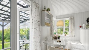 Interior claro com uma mesa de jantar, cadeiras e um banco. Há janelas com cortinas, uma luminária pendente e vista para o verde lá fora.