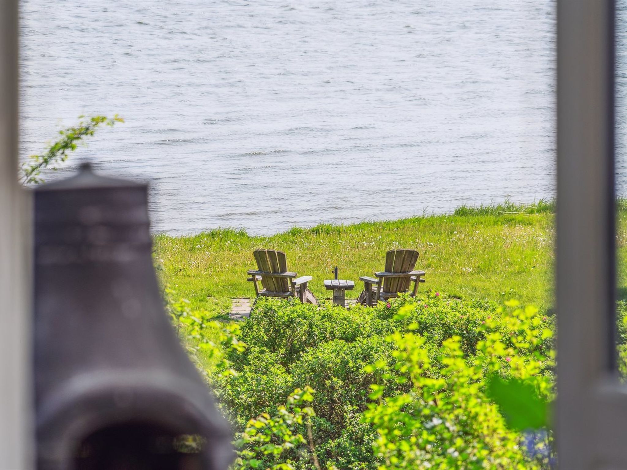 A lakeside view with three Adirondack chairs and a small table, surrounded by lush greenery.
