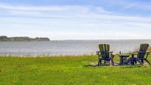 Vista para o lago com duas cadeiras Adirondack e uma pequena mesa, com vista para águas calmas e terra distante.