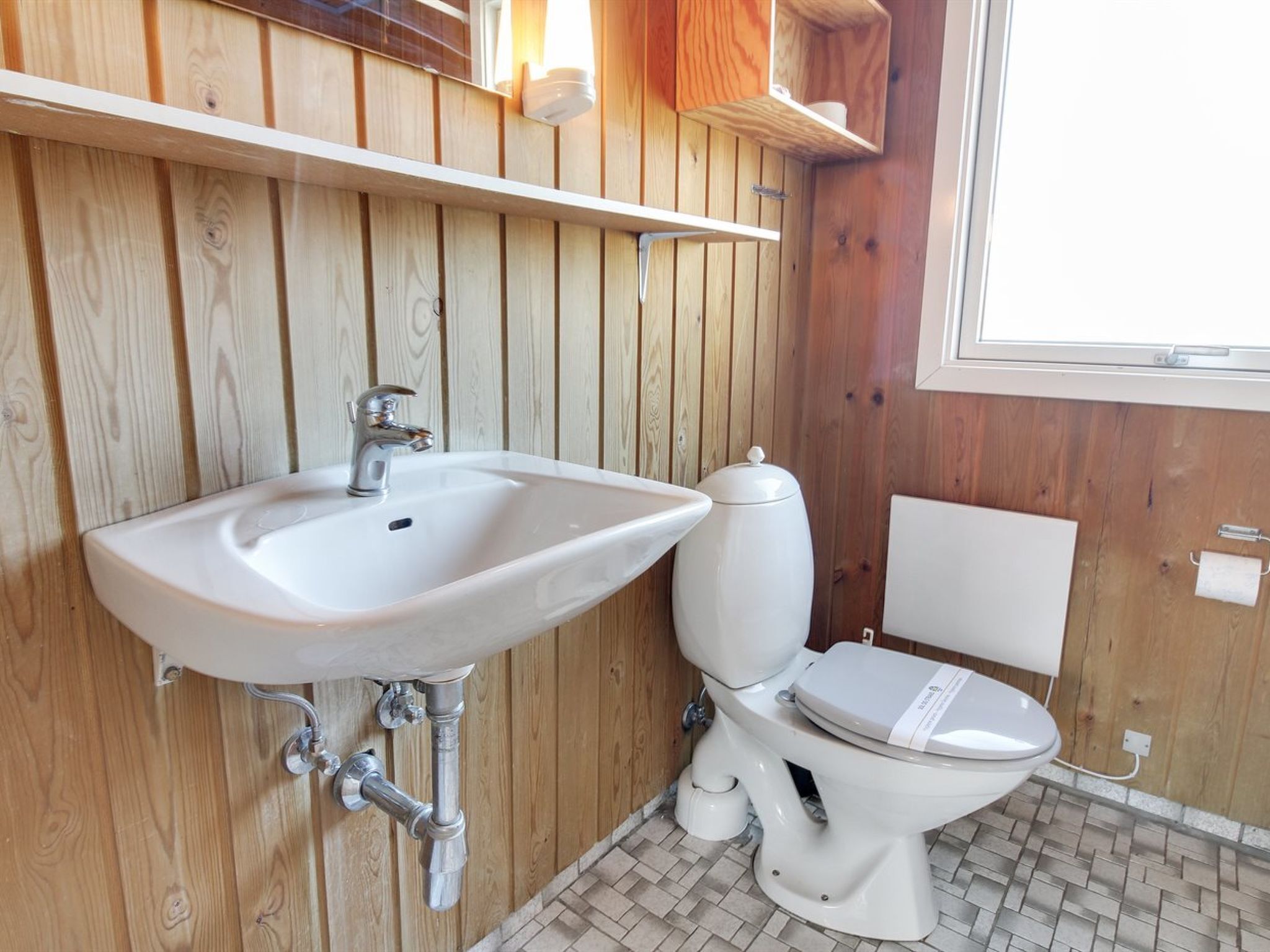 Un baño con pared de madera, un lavamanos blanco, un inodoro y una ventana.