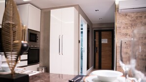 A modern kitchen with white cabinets, a microwave, and a stainless steel oven. There is a glass door leading to a bathroom, and a dining area with a table set for two. - MG Hill Hotel (Malatya)
