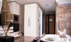 A modern kitchen with white cabinets, a microwave, and a stainless steel oven. There is a glass door leading to a bathroom, and a dining area with a table set for two.