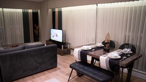 A modern living room with a sofa, a dining table set for two, and a television in the background. - MG Hill Hotel (Malatya)