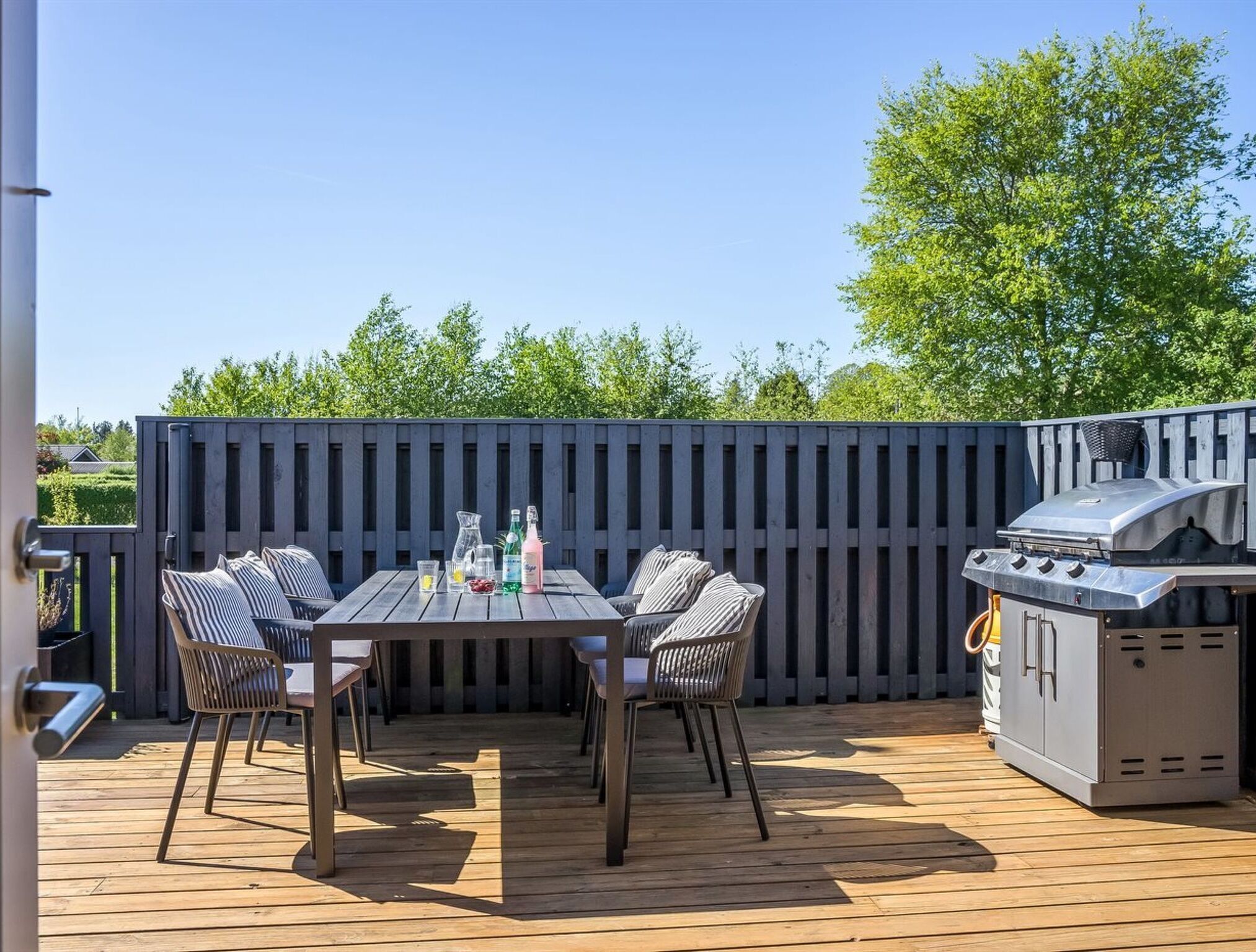 An outdoor patio with a wooden deck, a table with chairs, a grill, and a fence.