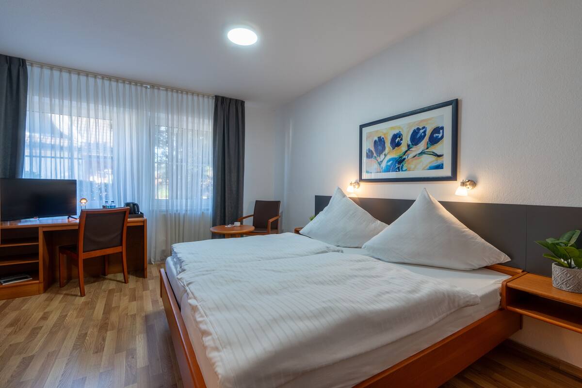 A hotel room with a bed, a desk with a chair, a television, and a painting on the wall.