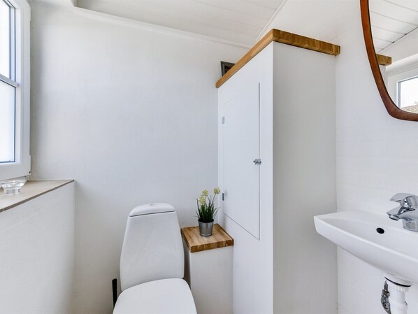A modern bathroom with a white toilet, a wooden shelf with a potted plant, a white cabinet, and a white sink with a mirror above it.