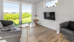 Ein modernes Wohnzimmer mit großem Fernseher, zwei Sofas, einem runden Couchtisch und großen Fenstern mit Blick auf Grünflächen.