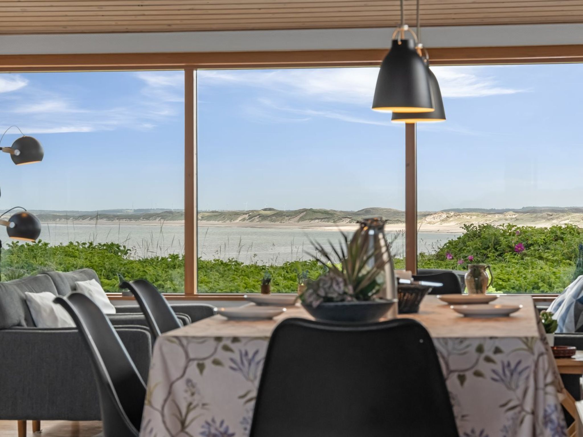 Un coin repas avec vue sur la mer, une table recouverte d’une nappe à motifs floraux et un luminaire suspendu noir.
