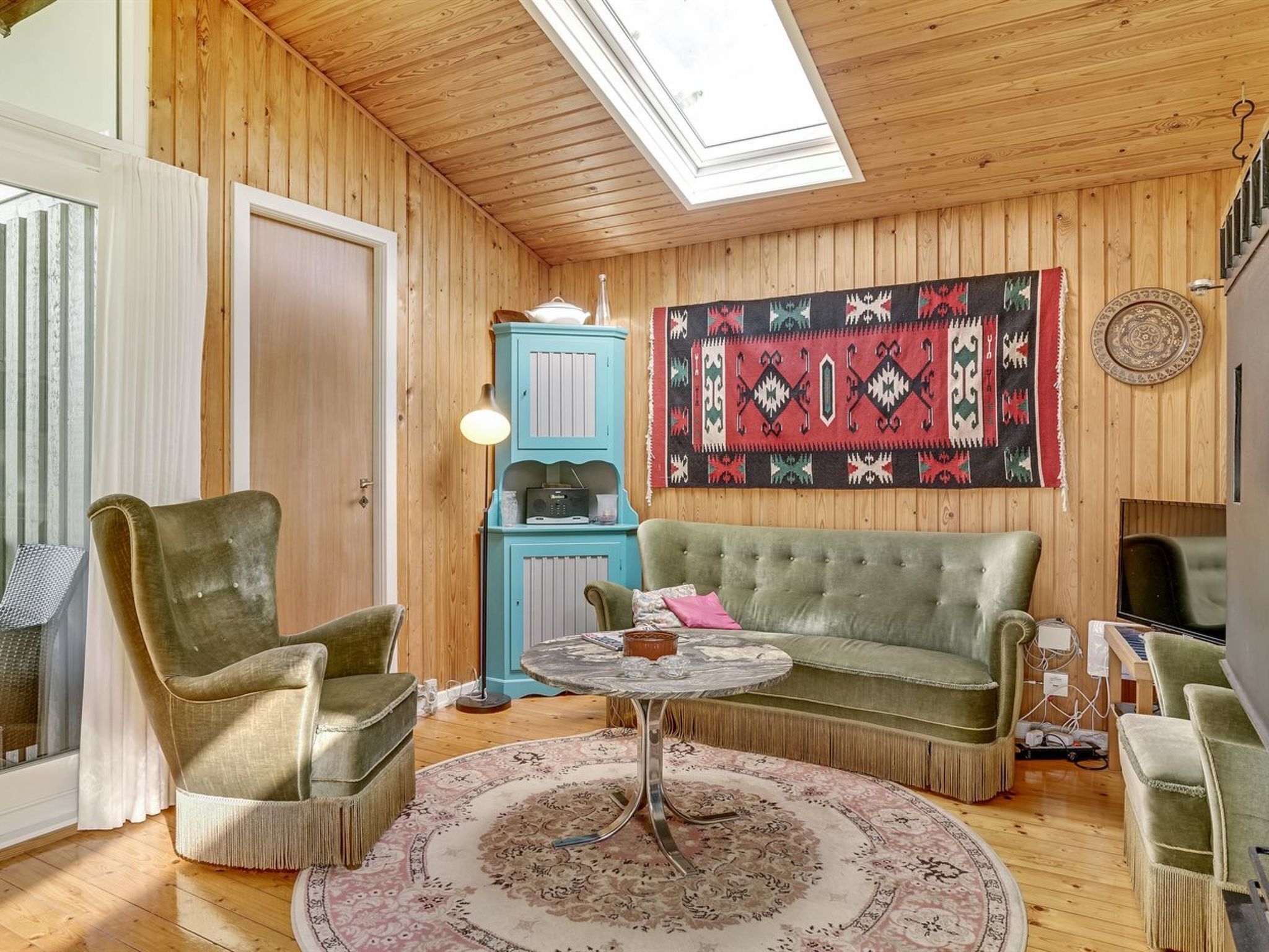 Een gezellige kamer met houten wanden, een groene bank, twee fauteuils, een klein tafeltje, een vloerkleed met een patroon, een blauwe kast met een radio, en een decoratieve wandversiering.