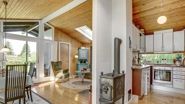 Une cuisine spacieuse dotée d’un plancher en bois, d’un poêle à bois central et d’une grande fenêtre donnant sur un espace verdoyant.