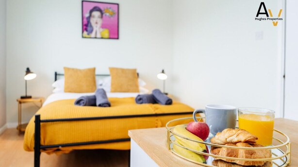 A neatly made bed with yellow bedding and two pillows, a bedside table with a tray of breakfast including fruit, croissants, and orange juice, and a framed picture on the wall. - Flexible 2-Bed House: Ideal for Business Trips (Northampton)
