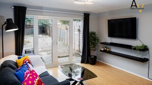 A modern living room with a sofa, a glass coffee table, a TV mounted on the wall, and a sliding glass door leading to an outdoor area. - Flexible 2-Bed House: Ideal for Business Trips (Northampton)