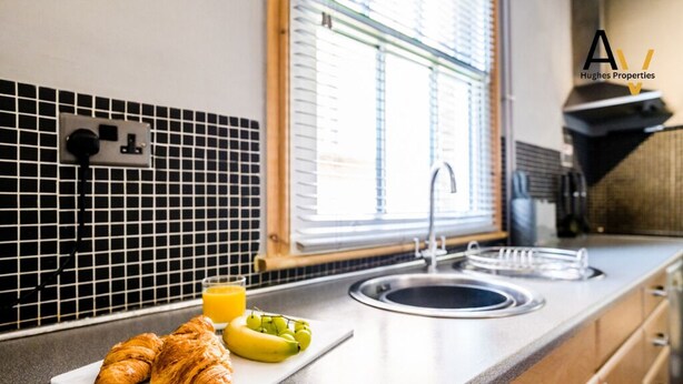 A kitchen with a black and white tiled backsplash, a window with blinds, a sink, and a countertop with croissants and bananas.