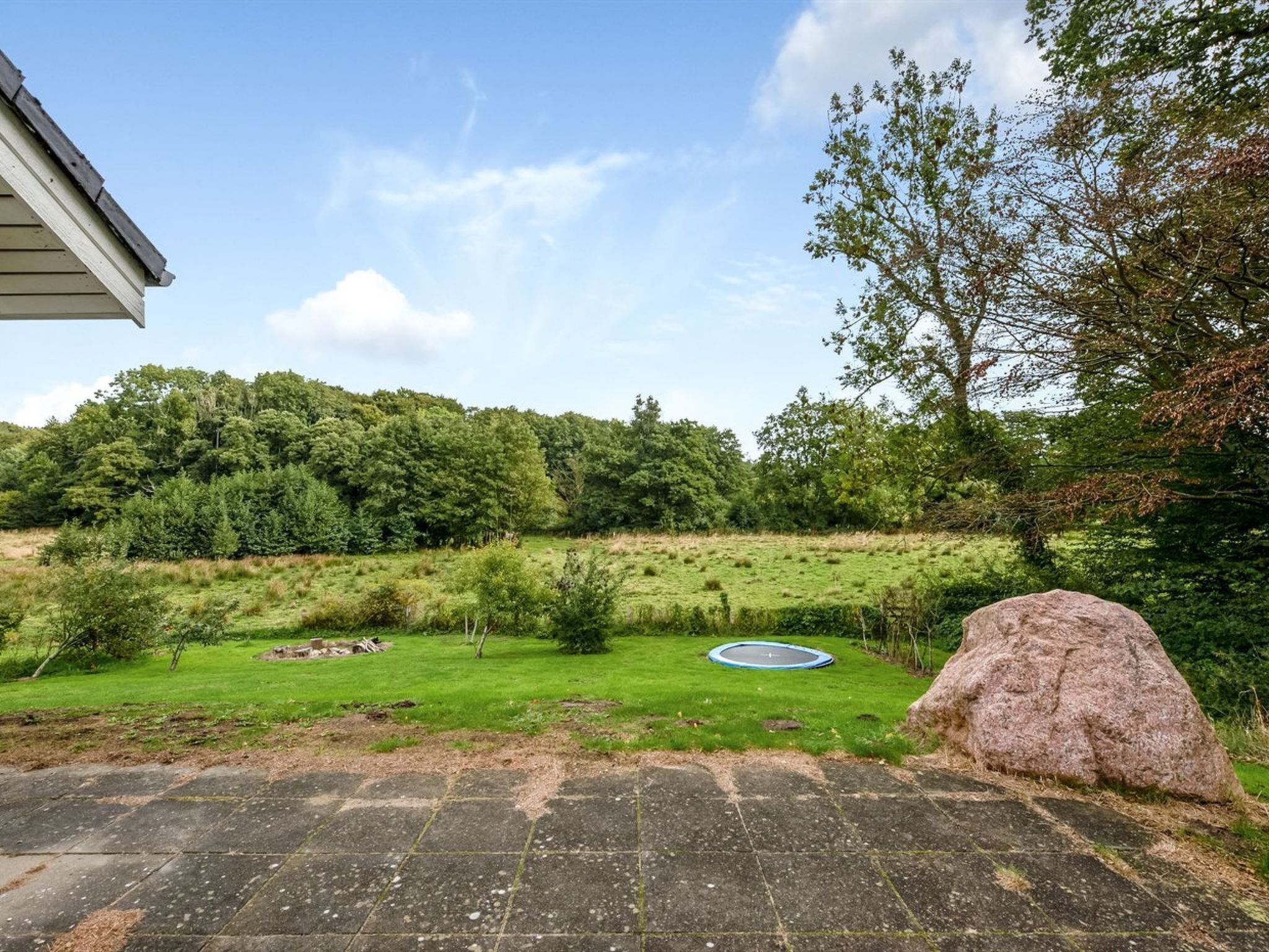 A backyard with a trampoline, a large rock, and a house with a sloped roof.