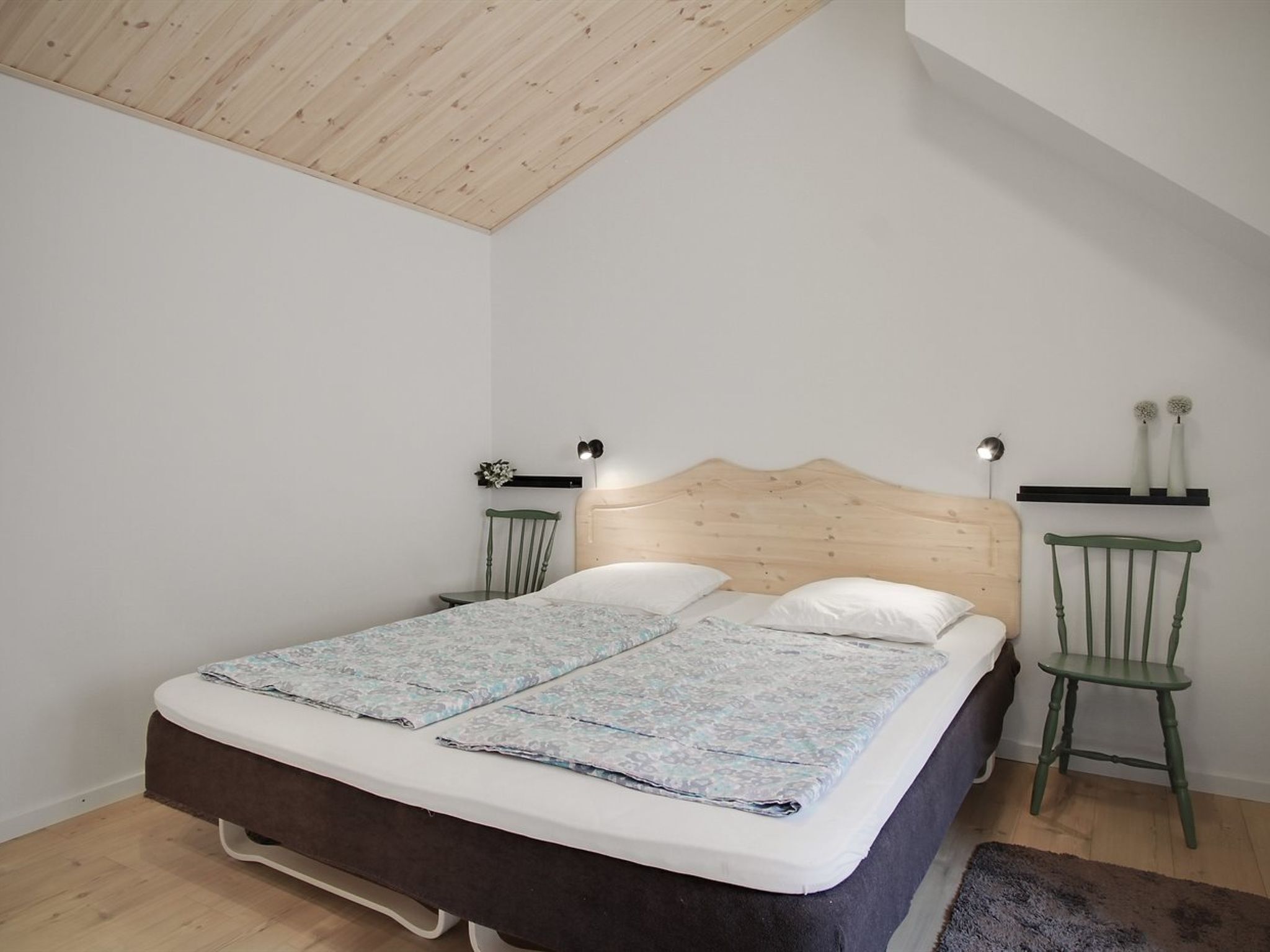 Une chambre avec une tête de lit en bois, un lit recouvert d’une couvre-lit à motifs floraux, deux tables de chevet avec des lampes et un fauteuil vert.
