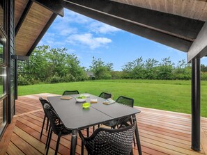 A covered outdoor patio with a table and chairs, a view of a grassy field, and a house in the background.