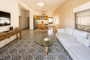 A modern living room with a glass coffee table, white sofa, patterned floor tiles, and a kitchen in the background.