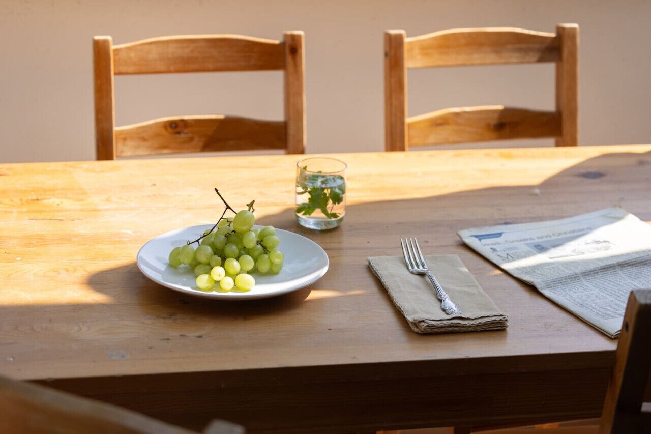 A wooden table with a plate of green grapes, a glass of water with a plant, a folded napkin, a fork, and a newspaper.